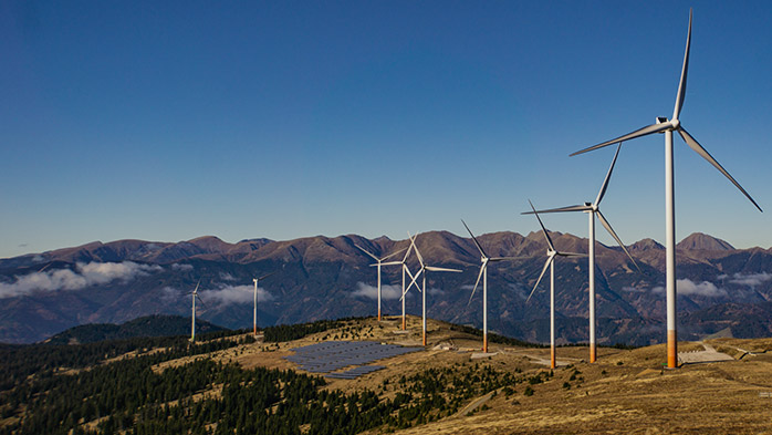 Wind turbines harness strong wind power on a grassy hill, with solar panels nearby, all set against a backdrop of mountains under a clear blue sky.