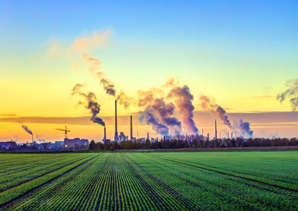 Green crop fields in neat rows extend under a bright blue and yellow sky as distant smokestacks emit dark smoke, depicting rebound power demand